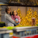 A parent and child in winter clothes marvel at a twinkling display of miniature buildings and trains