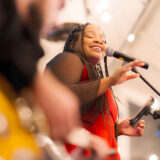 A person in a red dress sings while accompanied by a guitarist