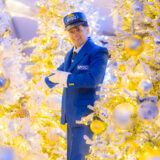 A person in a blue conductor's outfit stands among brightly lit white holiday trees