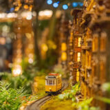 A model yellow trolley moves along toy train tracks, passing by warmly lit miniature homes