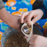 Two people work to place fragrant evergreen plant parts in a small cloth bag