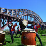 People on a lawn, using massive drums, play out a beat with a bridge in the background