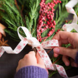 A person in a purple sweater ties a festive white ribbon around a bundle of green pine stems and red berries