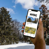 A person holds up a phone in front of a winter scene, showing an app with "NYBG" at the top