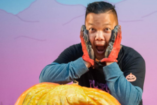 A person in red gloves makes a surprised face while posing with an orange pumpkin