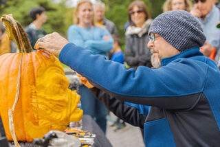 A person in a blue jacket and gray beanie carves a large orange pumpkin