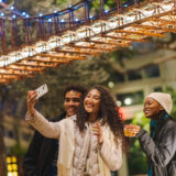 Three people pose for a selfie amid warmly lit models of bridges and buildings