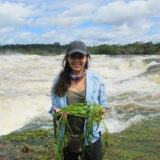 A girl with a black hat and blue shirt stands in front of the ocean and holds a bunch of kelp.