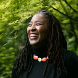 A person with long dark hair, wearing a black shirt and orange beaded necklace, poses under a green-leafed tree for a photo