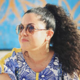 A person with long dark hair, wearing sunglasses and a blue and white shirt, poses in the sun for a photo