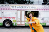 a person in a yellow coat and green oversized hat points to a double decker "Tea Around Town" bus parked on the street