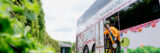 a person in a yellow coat and large hat is leaning out of a Tea Around Town double decker bus, with the New York Botanical Garden exterior sign in the background