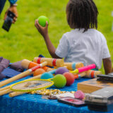 A child grabs a green ball from a table of outdoor lawn game equipment