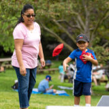 A child in a blue shirt and baseball hat throws red beanbags in a cornhole game while an older person in a pink shirt and jeans looks on.