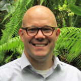 A person with a shaved head, dark glasses, and a white shirt poses outdoors for a photo