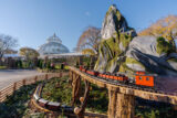 A red model train moves along a wooden bridge as it passes by a miniature mountain