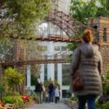 People walk along a path planted with green foliage, under and around models of city bridges made of plant parts