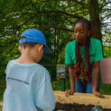 A young child green peers into a tub filled with soil while an older instructor in a green shirt looks on.