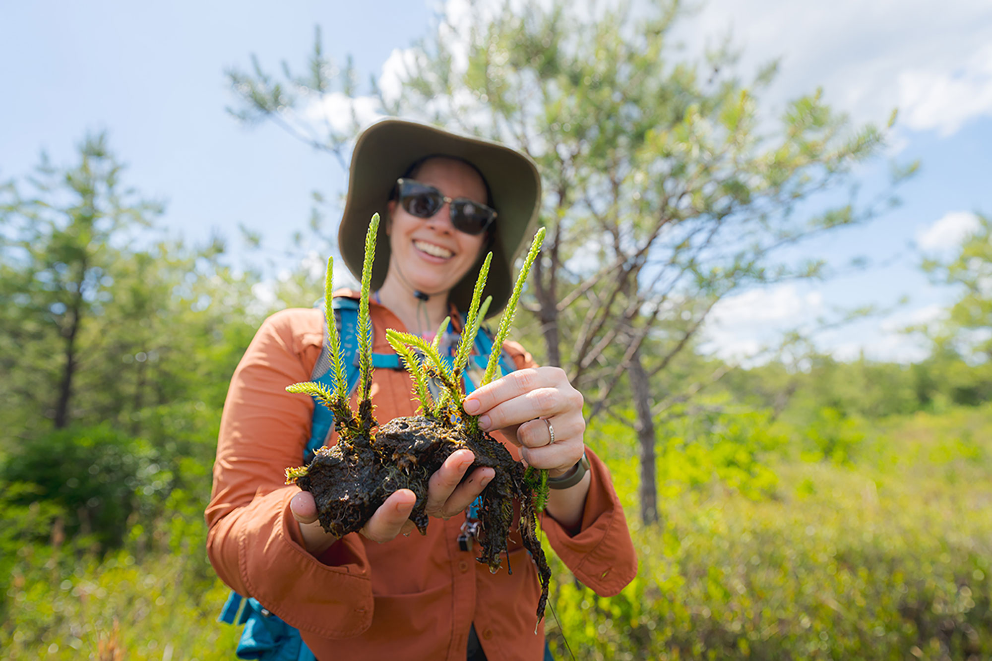 A woman in a hat and sunglasses holds up ferns that still have their roots and soil intact.