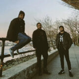 A group of three people in winter clothes pose for a photo while standing on the New York City coast