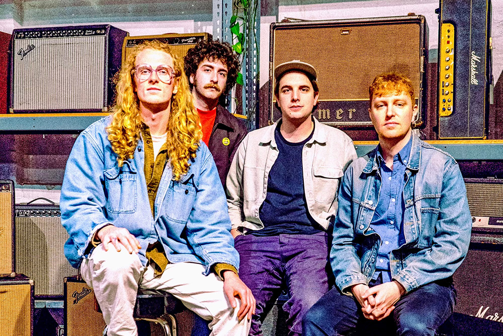 A group of four people pose for a photo in front of a collection of guitar amplifiers