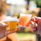 Two people clink small clear mugs full of beer in cheers