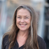 A person with long brown hair, wearing a black shirt, poses for a headshot