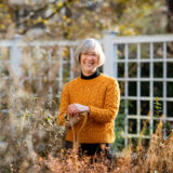 a person with short gray hair wears an orange sweater in a garden with fall plants