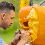 A person with short hair and a mustache carves a face into an orange pumpkin