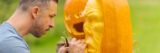 A person with short hair and a mustache carves a face into an orange pumpkin