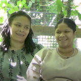 Two people in brown and green shirts speak while sitting in a sunny green garden