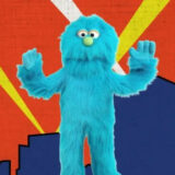 A blue puppet stands in front of a red, yellow, and blue background