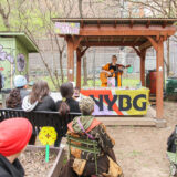 A person plays an acoustic guitar to a crowd while standing behind a table with a cloth reading "NYBG"