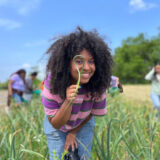 A person in a pink and purple sweater looks through a hoop of plant material while posing for a photo in a green field