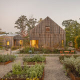 A soft yellow light can be seen in a wood paneled house. In the backyard, there are tables and chairs set up, surrounded by green trees.