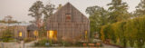 A soft yellow light can be seen in a wood paneled house. In the backyard, there are tables and chairs set up, surrounded by green trees.