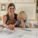 A woman with dark hair in brown overalls sits next to a blond child and draws on a blueprint for a landscape design.