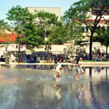 three children run on the concrete that is covered in water; there are adults sitting on benches in the background