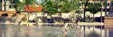 three children run on the concrete that is covered in water; there are adults sitting on benches in the background