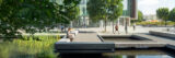 a person is sitting on the ledge of a walkway over a pond with tall green grass growing out of it. A glass building is in the background, and other people are walking in front of it.