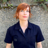 A woman with red hair and bangs wearing a dark blue collared shirt looks at the camera with a soft smile.