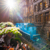 A blue model train zips past miniature buildings and white flowers as the sun shines from above