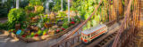 A model trolley in red and white travels along a sunny miniature bridge made of plant parts