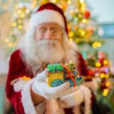 A person dressed as Santa, in red and white, offers a colorful train Christmas ornament