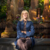 A person with long blonde hair and a blue jacket poses for a photo before a vignette of miniature buildings