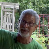 A person in a green shirt and glasses speaks from an outdoor garden