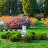 An armillary sphere sits among green shrubs in a colorful garden