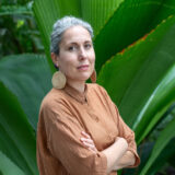 A person in a brown shirt, with large golden disc earrings, poses for a photo in front of a large, green tropical plant