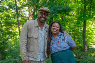 A man with a brown hat with the letters H C, glasses and a striped button down shirt stands next to a woman in a striped shirt with ladybugs and green pants.