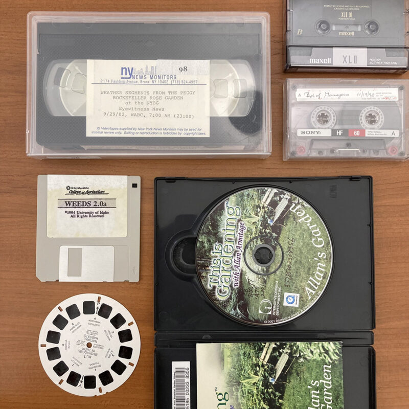 An arrangement of VHS tapes, floppy discs, CDs, cassette tapes, and ViewMaster discs on a wooden table